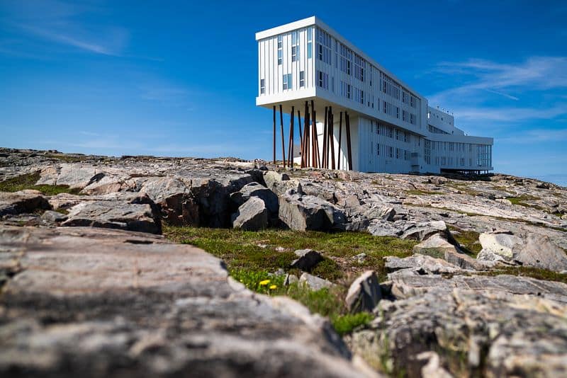 Bild von Fogo Island in Kanada: Ein modernes, weißes Gebäude mit vielen Fenstern steht auf hohen, schlanken Stelzen über einem felsigen Küstenabschnitt. Der Himmel ist klar und blau, und der Boden ist mit großen Steinen und etwas Gras bedeckt, während ein paar gelbe Blumen im Vordergrund sichtbar sind.
