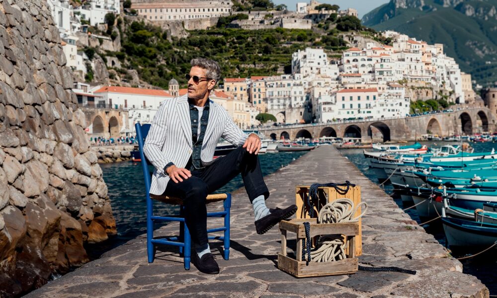 Mann im Hafen Amalfi Stilvoller Mann mit FALKE Socken sitzt auf einem Stuhl am Hafen von Amalfi, Fischerboote und weiße Häuserfassaden im Hintergrund