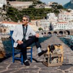 Mann im Hafen Amalfi Stilvoller Mann mit FALKE Socken sitzt auf einem Stuhl am Hafen von Amalfi, Fischerboote und weiße Häuserfassaden im Hintergrund