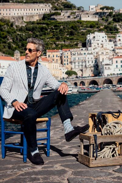 Mann im Hafen Amalfi Stilvoller Mann mit FALKE Socken sitzt auf einem Stuhl am Hafen von Amalfi, Fischerboote und weiße Häuserfassaden im Hintergrund