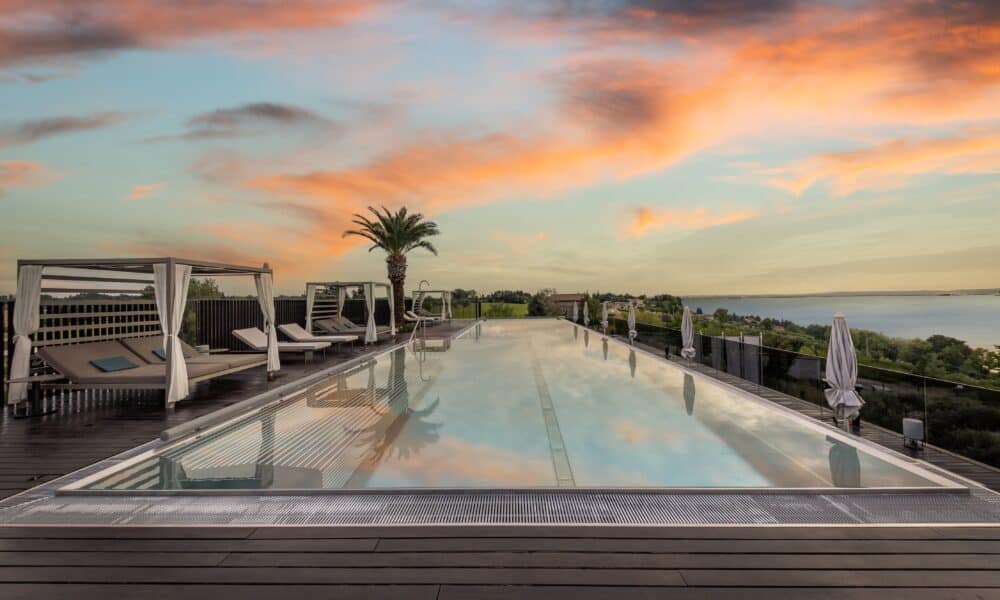 Infinity-Pool des Quellenhof Luxury Resort Lazise bei Sonnenuntergang mit Blick auf den Gardasee, flankiert von eleganten Daybed-Pavillons und einer Palme unter dramatisch gefärbtem Abendhimmel.