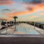 Infinity-Pool des Quellenhof Luxury Resort Lazise bei Sonnenuntergang mit Blick auf den Gardasee, flankiert von eleganten Daybed-Pavillons und einer Palme unter dramatisch gefärbtem Abendhimmel.