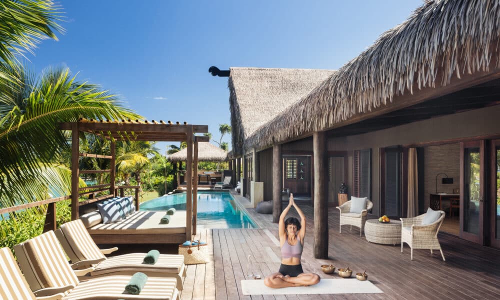 Frau bei einer Yoga-Übung auf der privaten Holzterrasse einer Pool-Villa im Six Senses Fiji, umgeben von Palmen, Strohdach-Architektur und Lounge-Liegen