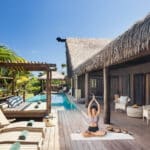 Frau bei einer Yoga-Übung auf der privaten Holzterrasse einer Pool-Villa im Six Senses Fiji, umgeben von Palmen, Strohdach-Architektur und Lounge-Liegen