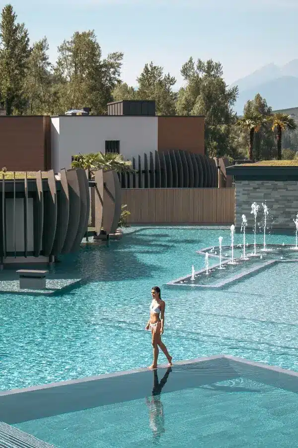 Schönste Hotelpools 2026: 6 spektakuläre Luxus-Pools für den Sommer Vor der Kulisse der Südtiroler Alpen flaniert eine Frau am kristallklaren Wasser der Quellenhof See Lodge zwischen moderner Architektur und sprudelnden Fontänen.