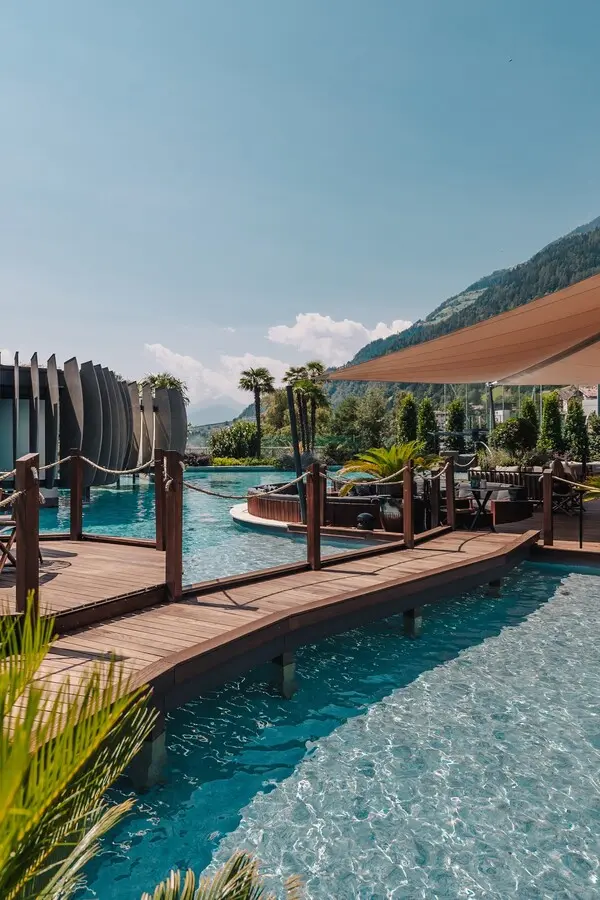 Schönste Hotelpools 2026: 6 spektakuläre Luxus-Pools für den Sommer Hölzerne Stege führen über die weitläufige Wasserlandschaft der Quellenhof See Lodge und verbinden modernes Design mit der alpinen Kulisse des Südtiroler Passeiertals.